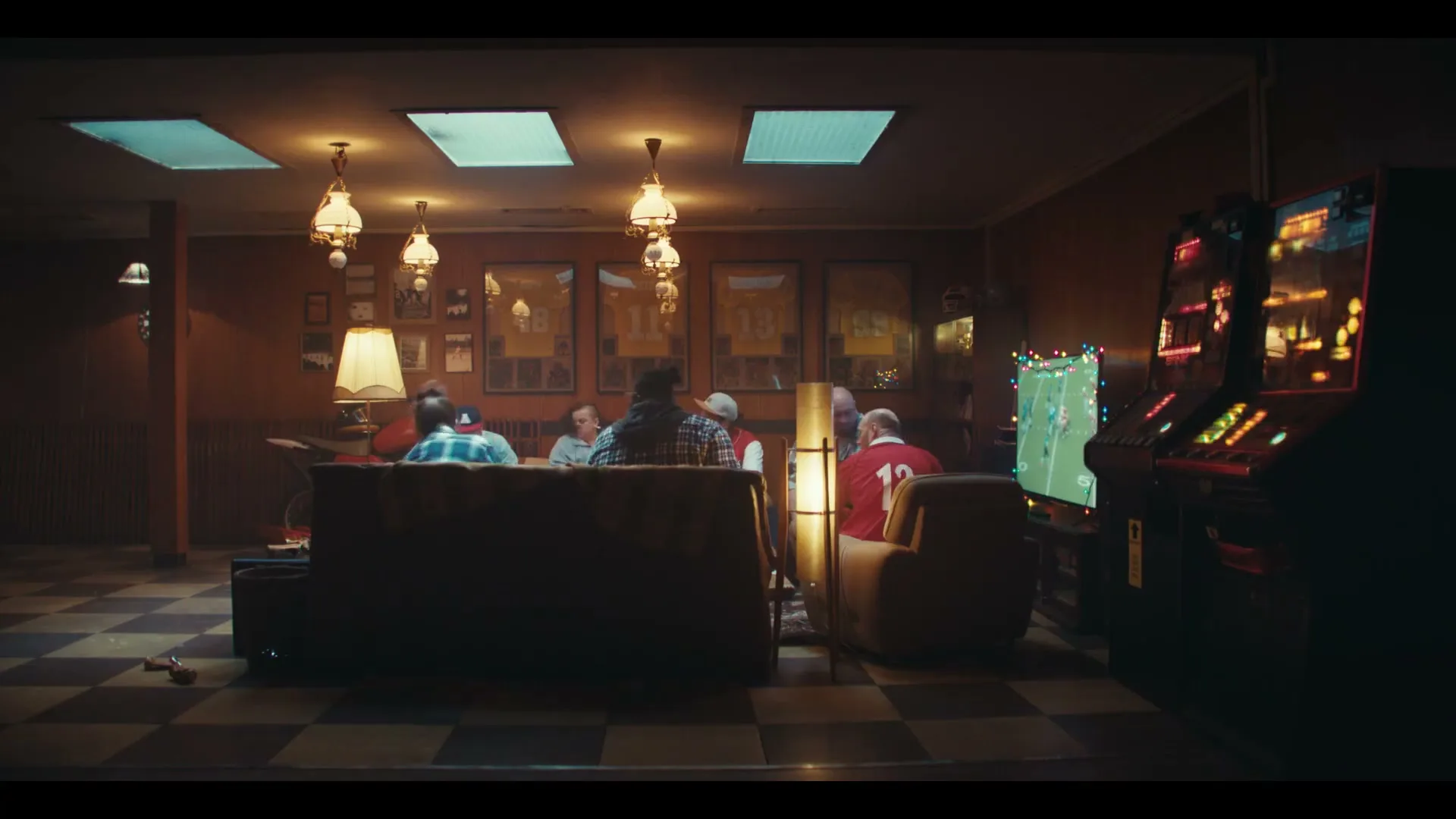 A group of men watch football on TV in a dimly lit, wood-paneled basement with arcade games and sports memorabilia.