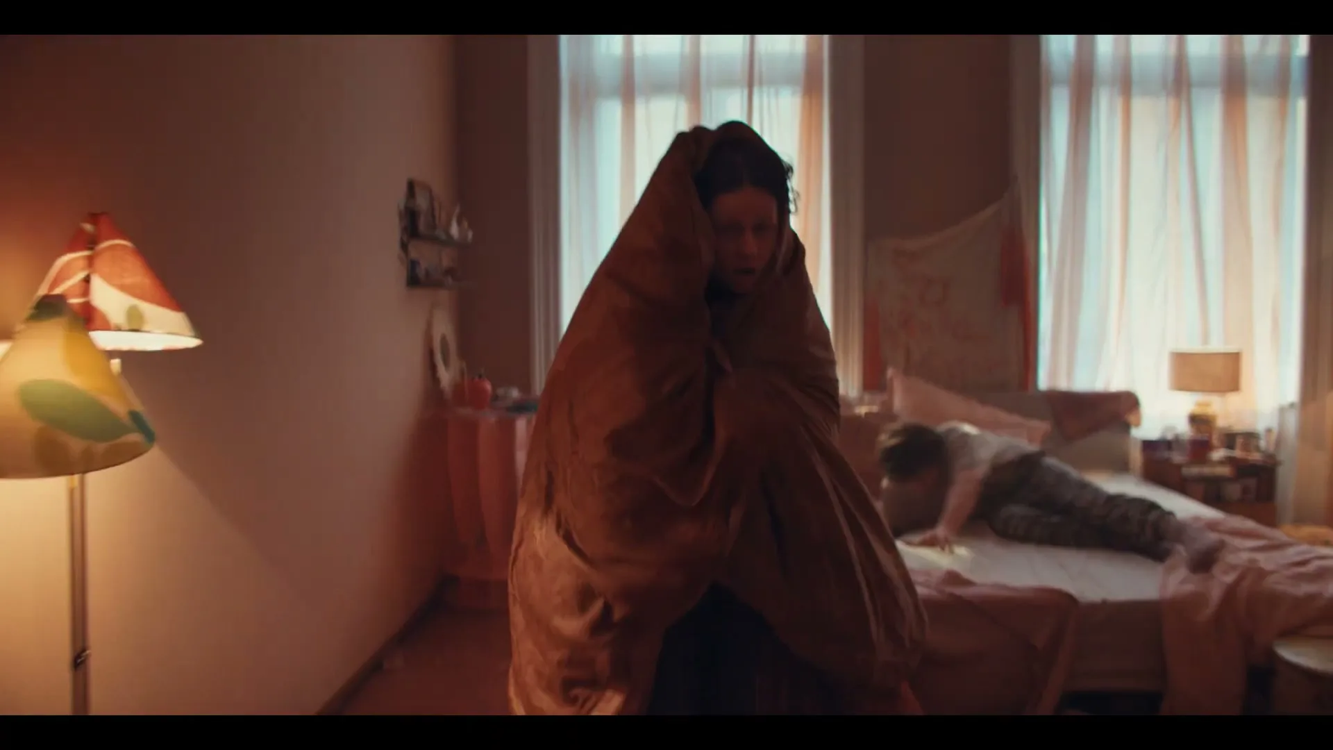 A young woman, wrapped in a brown blanket, stands in a messy bedroom while another person is on the bed.