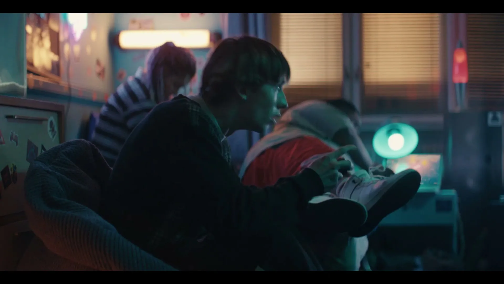 Three young adults are seen in a dimly lit room, with one person in the foreground playing a video game while holding a controller.
