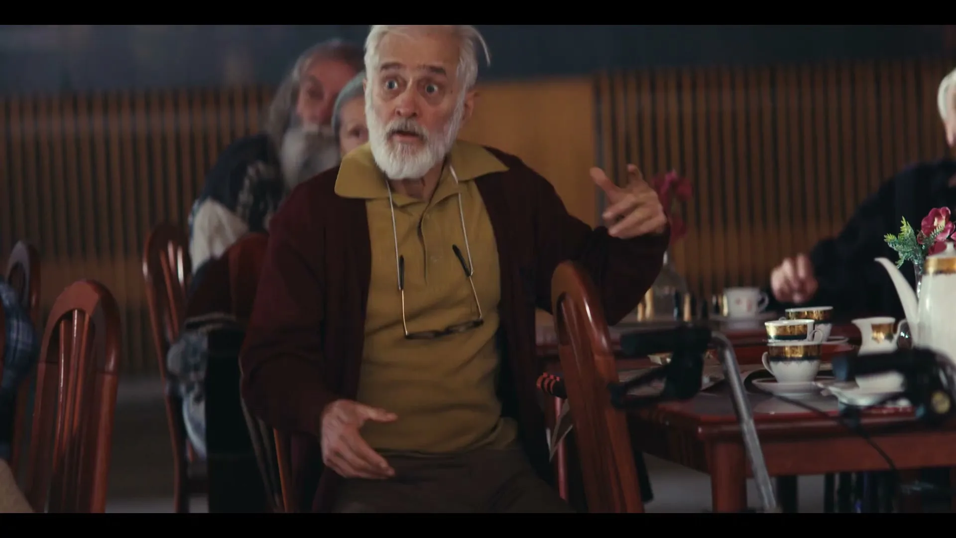 A surprised elderly man with a white beard and glasses around his neck sits at a table in a dimly lit room, looking wide-eyed.