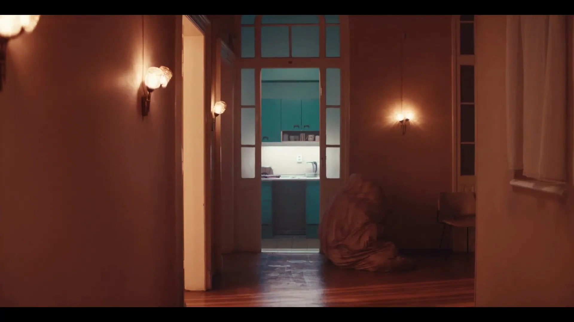 A young man and woman, both in pajamas, emerge from a doorway into a dimly lit hallway, the woman wrapped in a blanket.