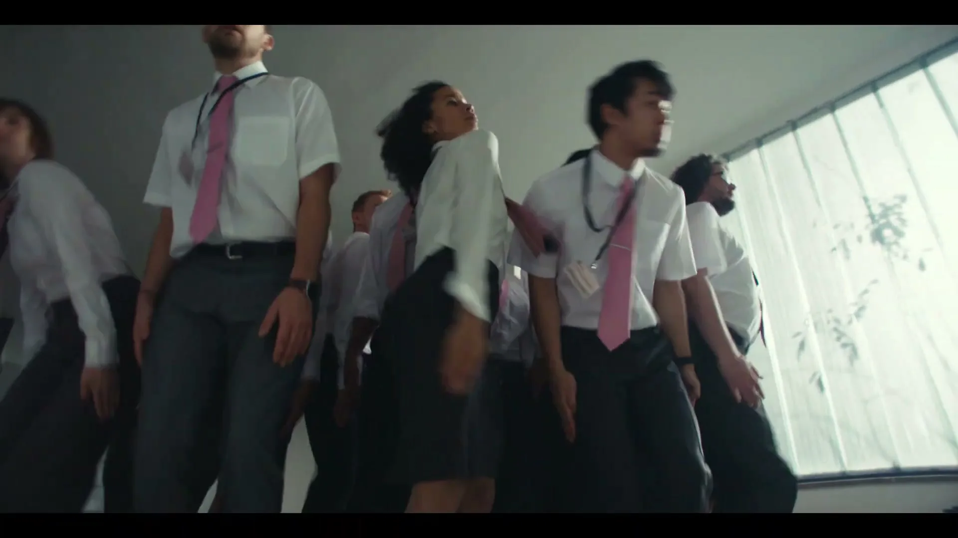 A diverse group of office workers in white shirts and pink ties are dancing in an office building with large windows.