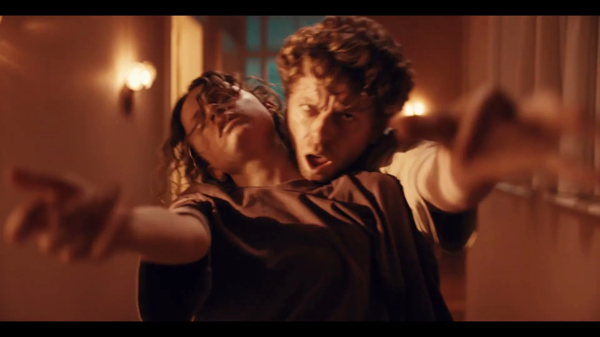 A man and a woman, both with disheveled hair and wearing dark t-shirts, are intertwined and reaching out with intense, open-mouthed expressions in a dimly lit hallway.
