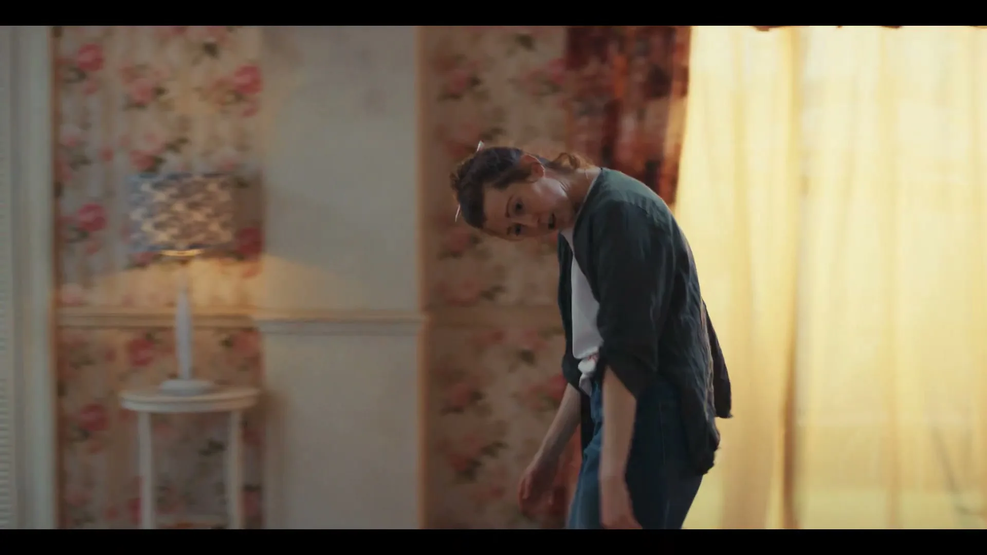 A woman with hair curlers and a dark cardigan dances energetically in a room with floral wallpaper and a lamp.