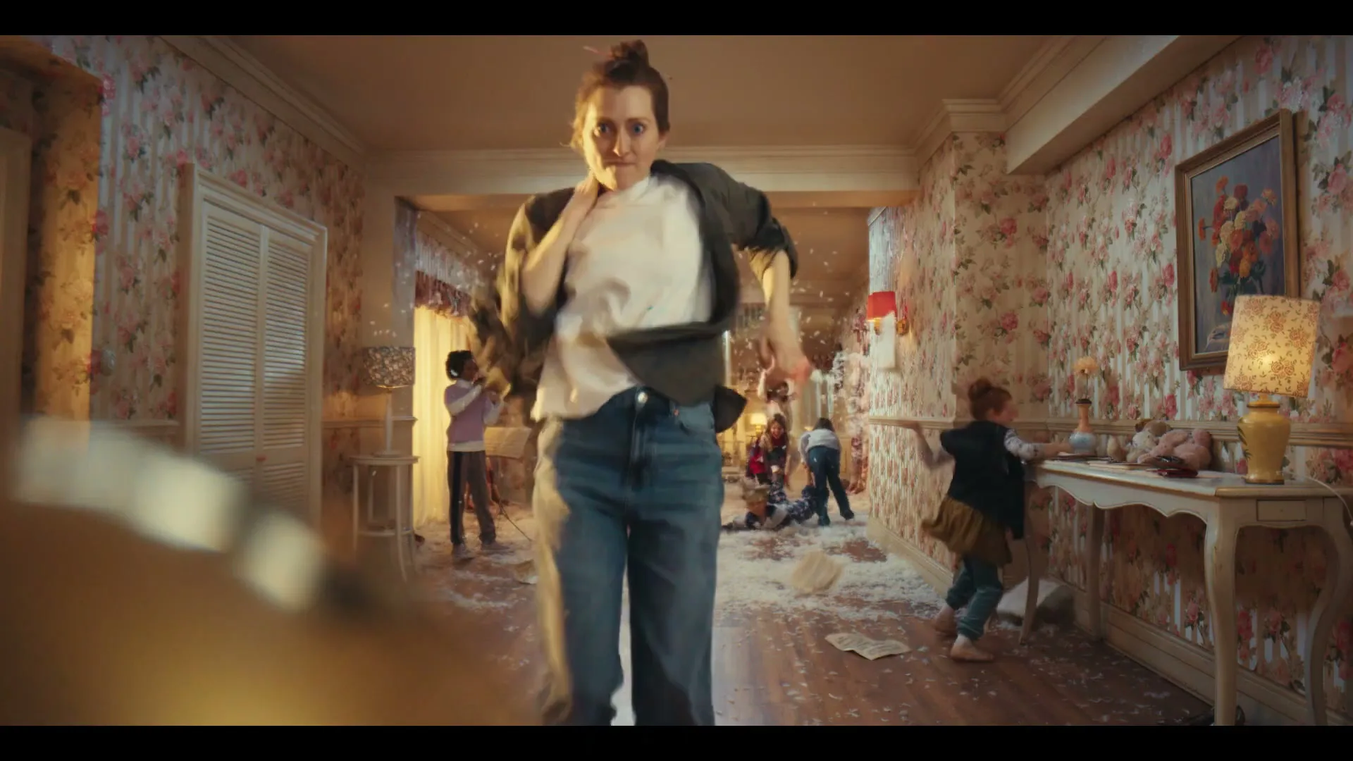 A woman in a messy house, covered in feathers, runs towards the camera with a frantic expression while children play in the background.
