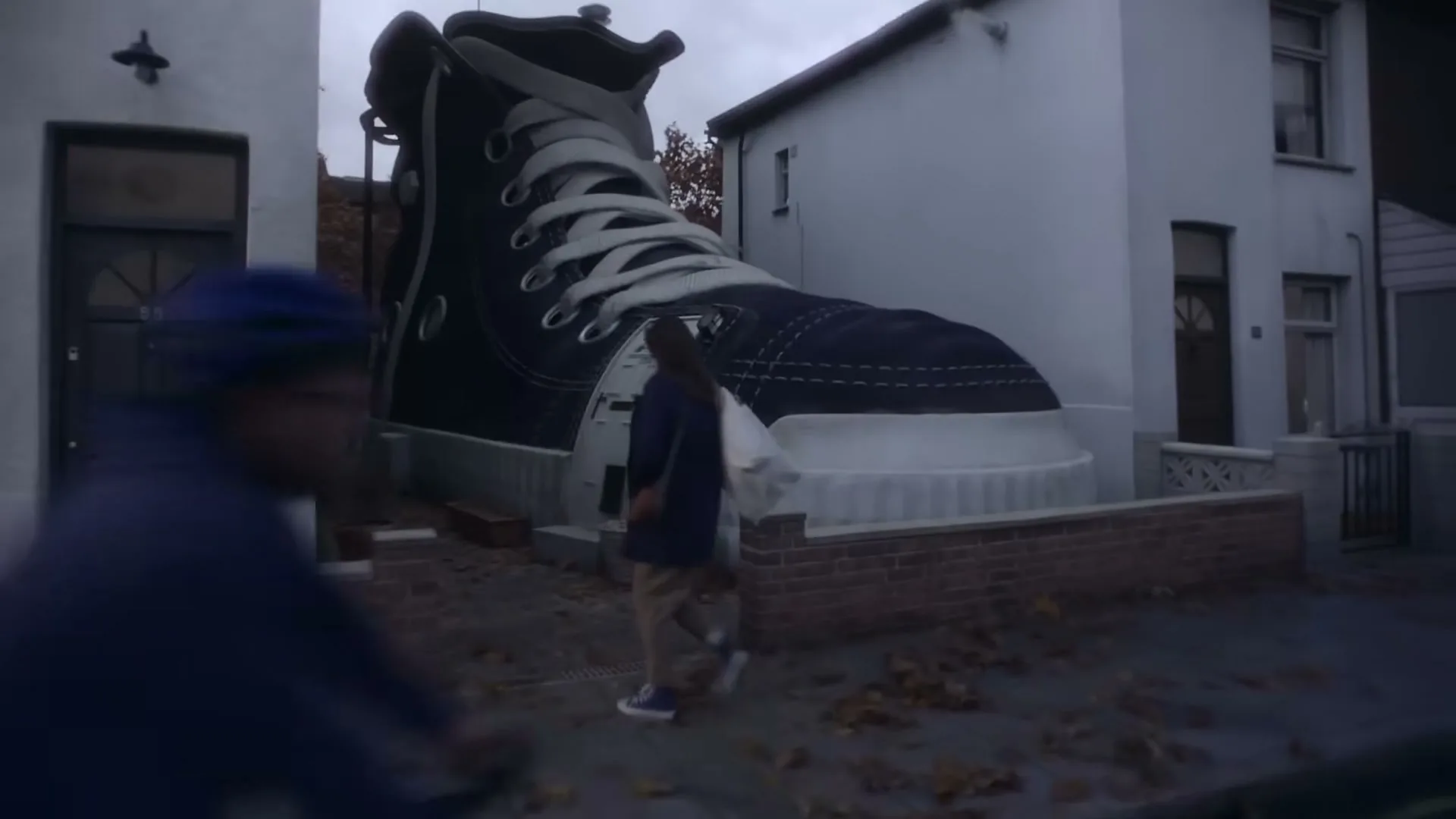 A woman walks down a suburban street, looking at her phone, then a giant shoe appears between two houses as she walks past.