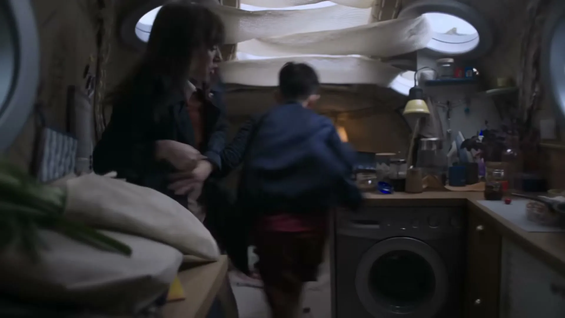 A woman and a child move through a small, cluttered living space with a kitchen area, as another person sits in the background.
