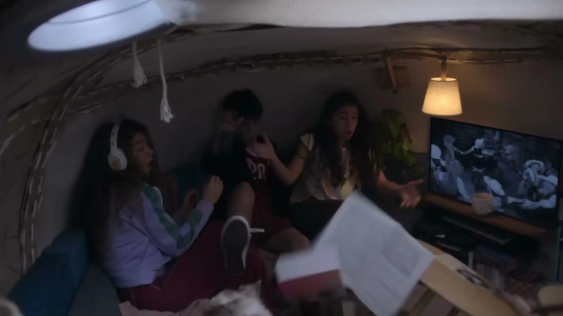 Three teens are startled while watching a black and white movie in a blanket fort at night, causing papers to fly.