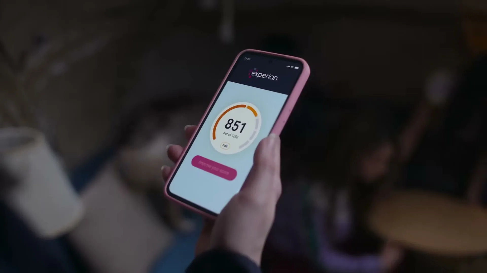 A person holds a pink smartphone displaying the Experian app with a credit score increasing rapidly, in a dimly lit interior.