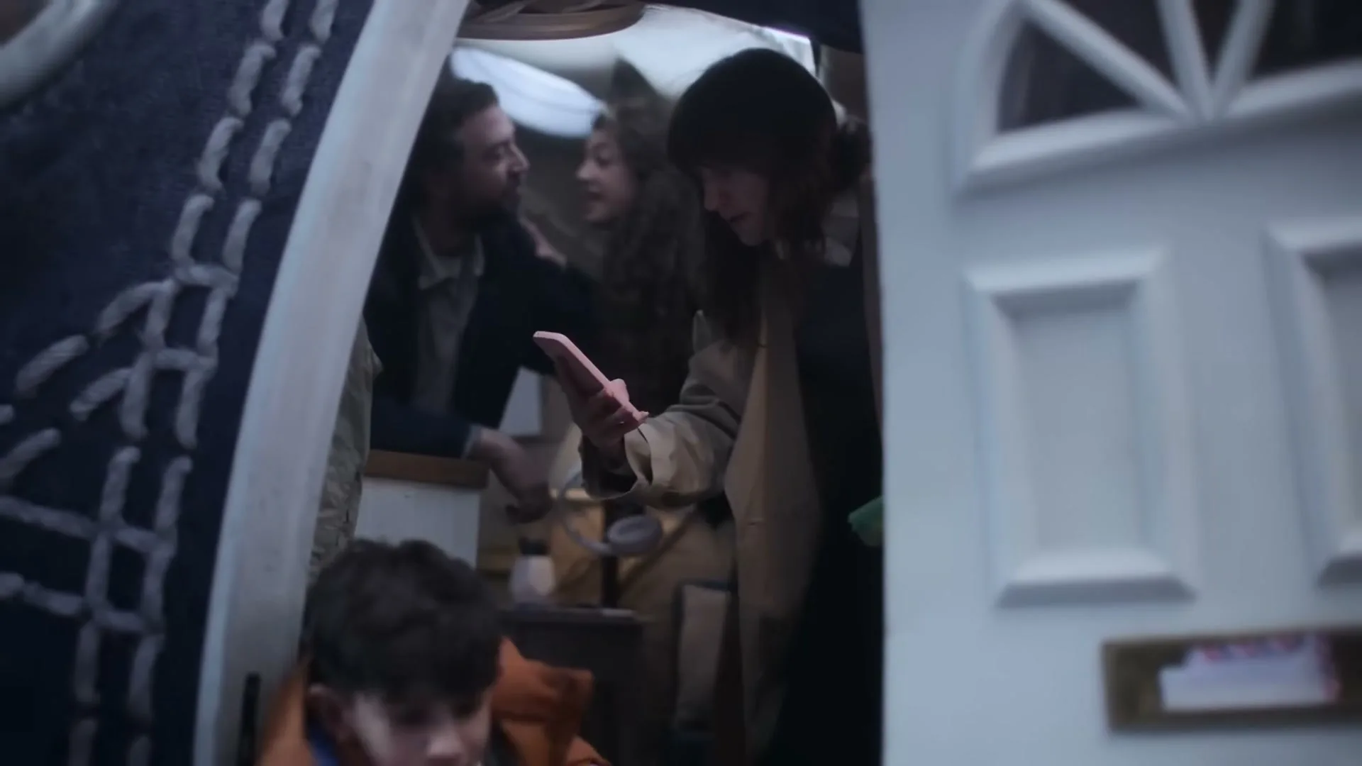 A family of four, including a mother in a trench coat and a young boy in an orange jacket, are seen in a medium shot, rushing out of their front door.