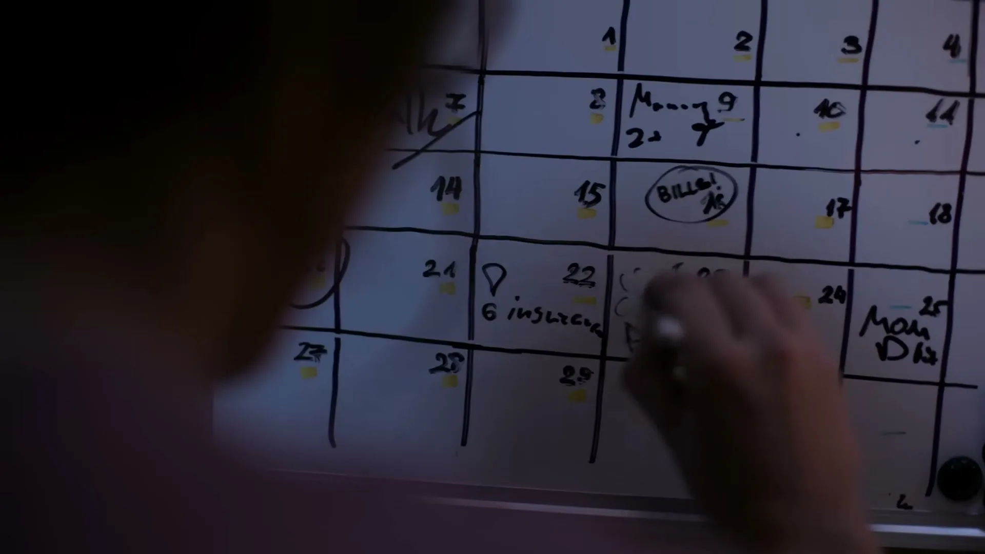 A person's hand writes on a whiteboard calendar with various notes and dates, including "BILLS!" circled on the 16th.