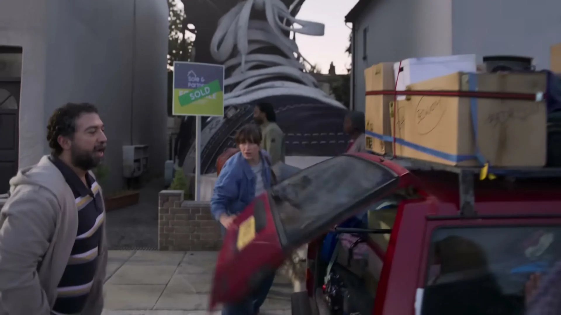 A family packs a red car with boxes and belongings in front of a house with a "Sold" sign and a giant shoe sculpture.
