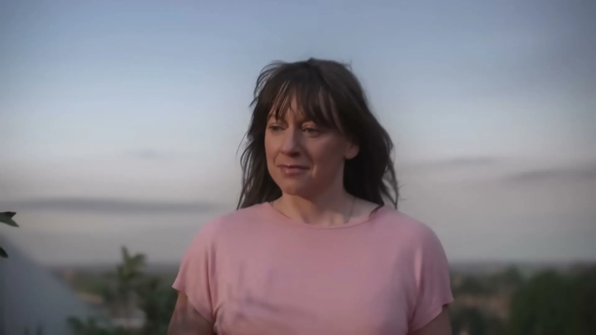 A woman with dark hair and a pink shirt smiles softly, looking down and to the right, with a blurred cityscape and sky in the background.