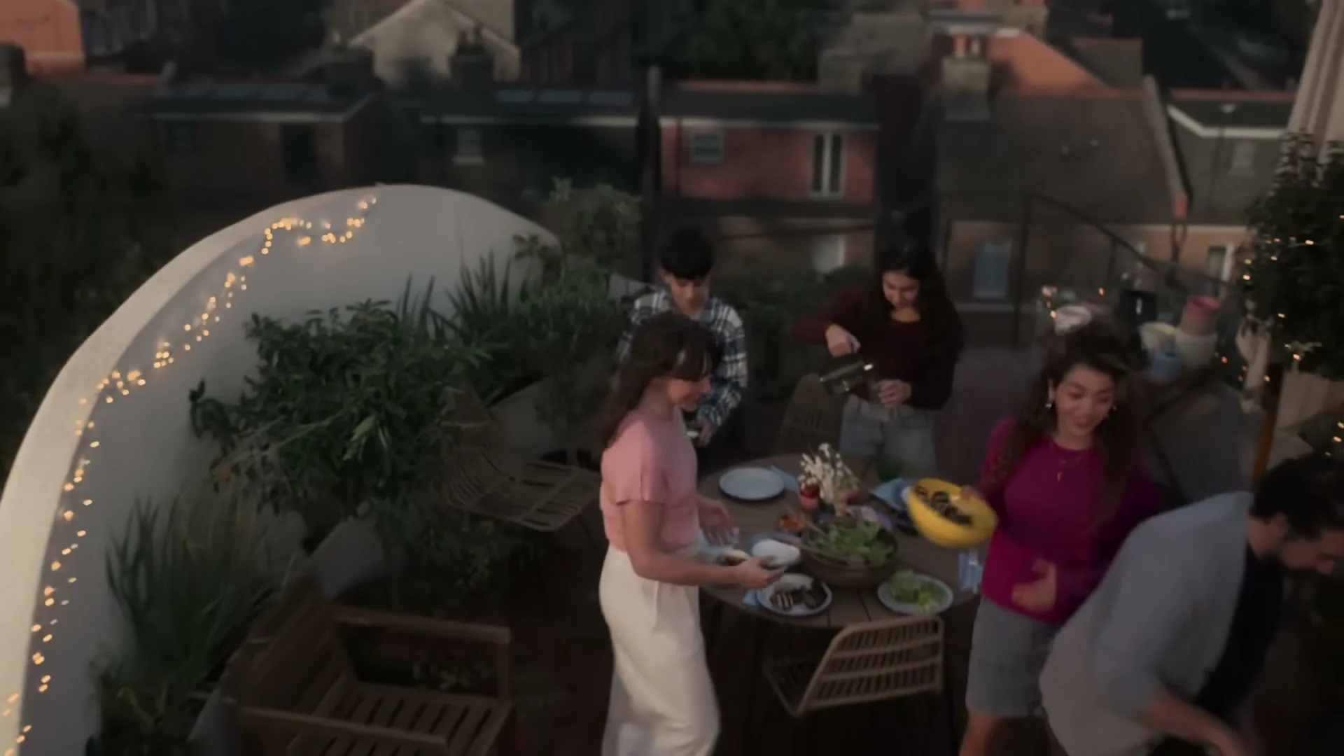 A diverse group of young adults and a dog enjoy a rooftop barbecue at dusk, with city buildings in the background.