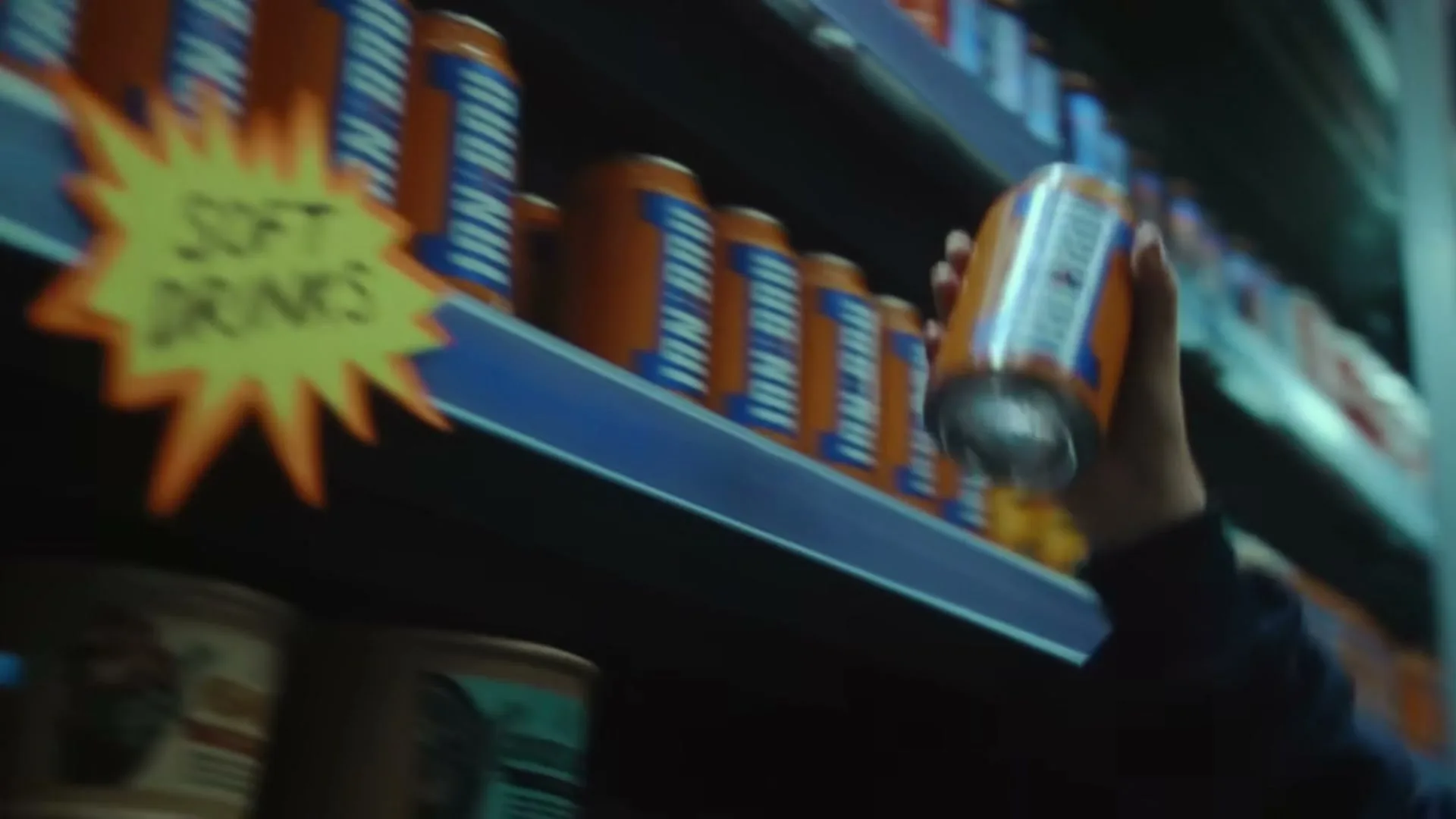 A young boy with red hair and freckles picks up a can of IRN-BRU from a supermarket shelf, looking confused by a "Soft Drinks" sign.