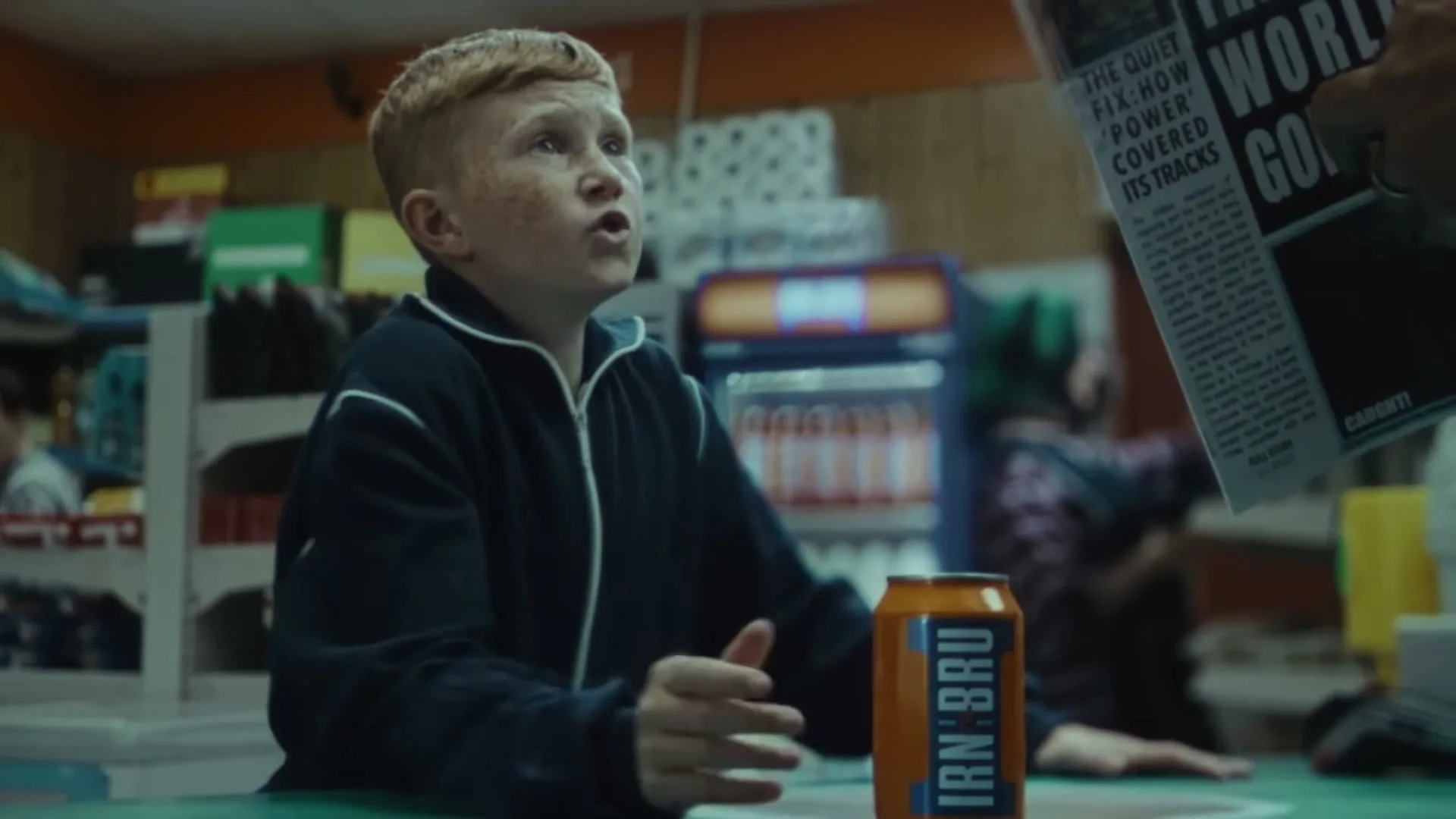 A young boy with red hair and freckles stands at a convenience store counter, placing an IRN-BRU can down as he looks up at someone off-screen.