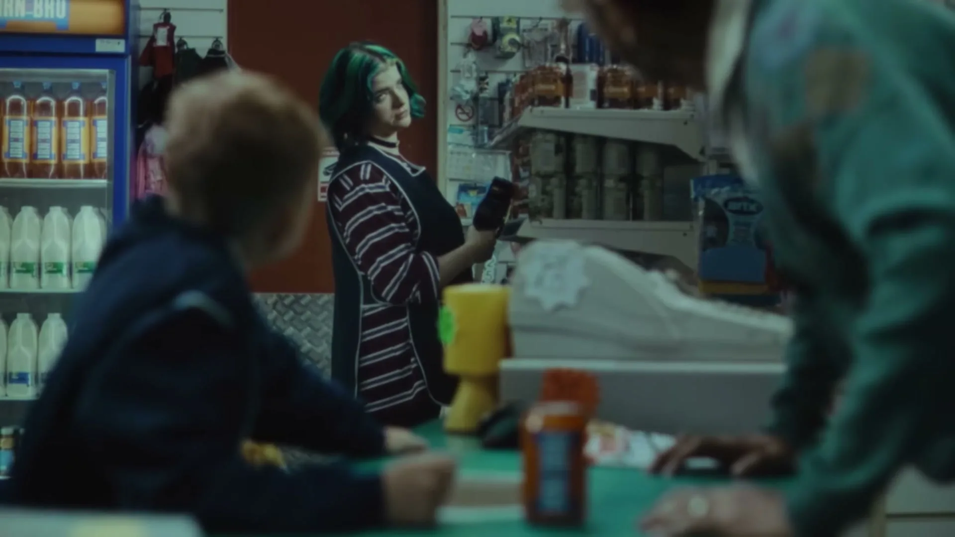 A young woman with green hair, wearing a striped apron, stands behind a counter in a convenience store, looking at a phone while a child and an adult stand in front of her.