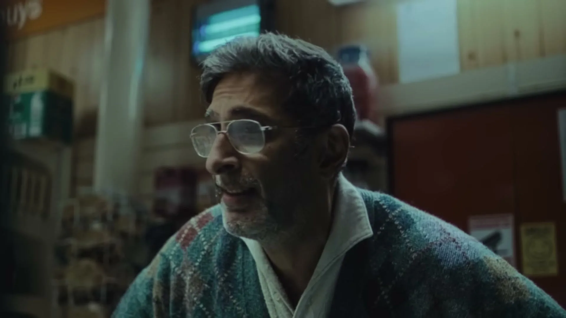 A middle-aged man with glasses and a patterned cardigan looks around a dimly lit convenience store with a concerned expression.