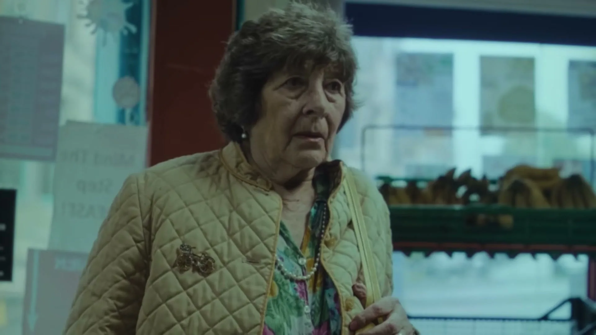 An elderly woman with short brown hair and a quilted jacket looks surprised and then turns to her right in a brightly lit store.