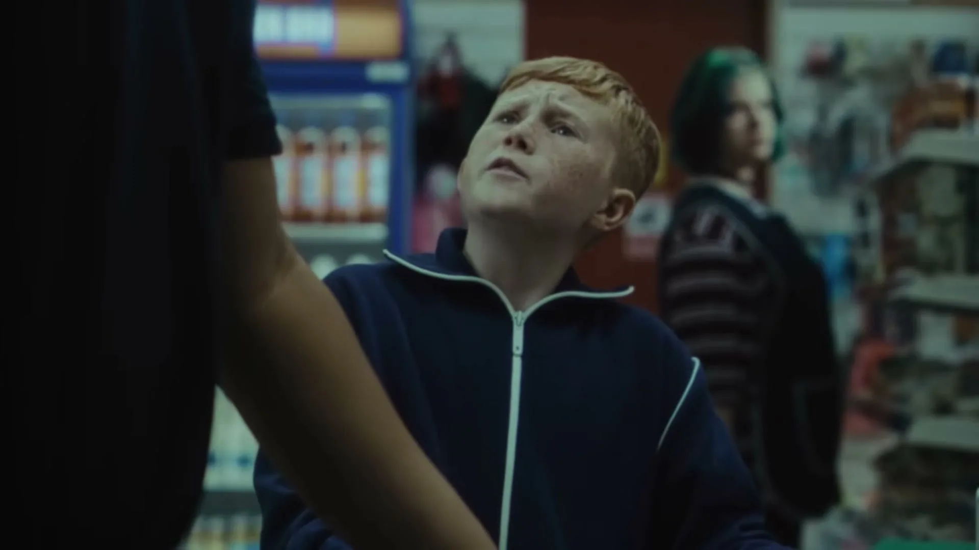 A young boy with red hair and freckles looks up at an unseen person, making a face in a convenience store.