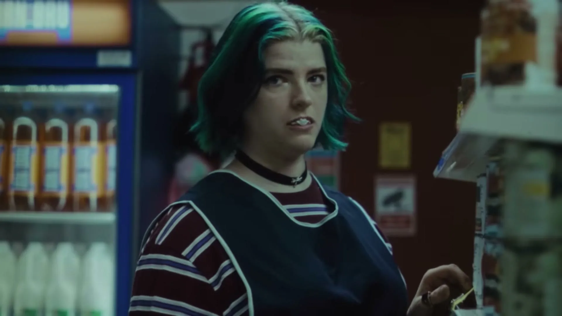 A young woman with green and blonde hair, wearing a striped shirt and apron, chews gum and looks around a convenience store.