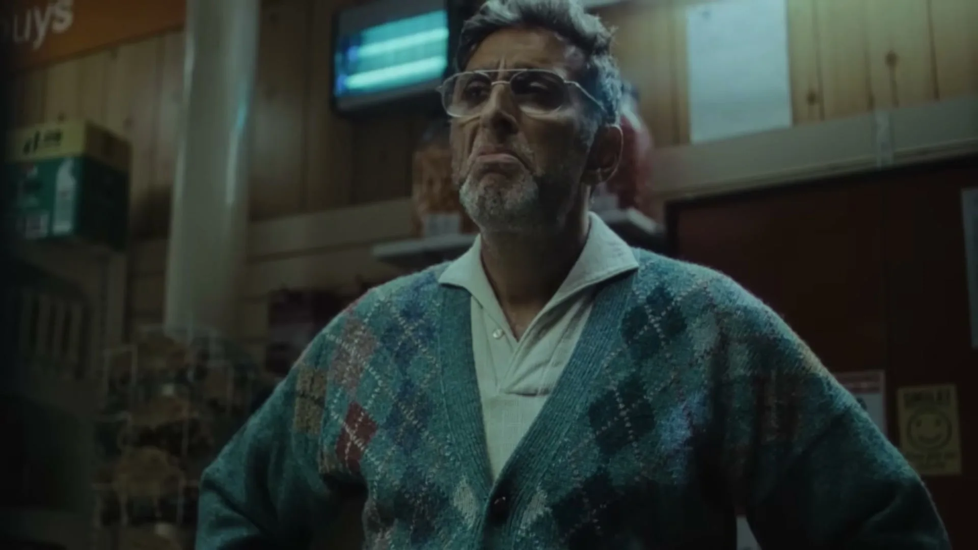 A man with glasses and a patterned cardigan stands with hands on hips, looking up in a dimly lit convenience store.