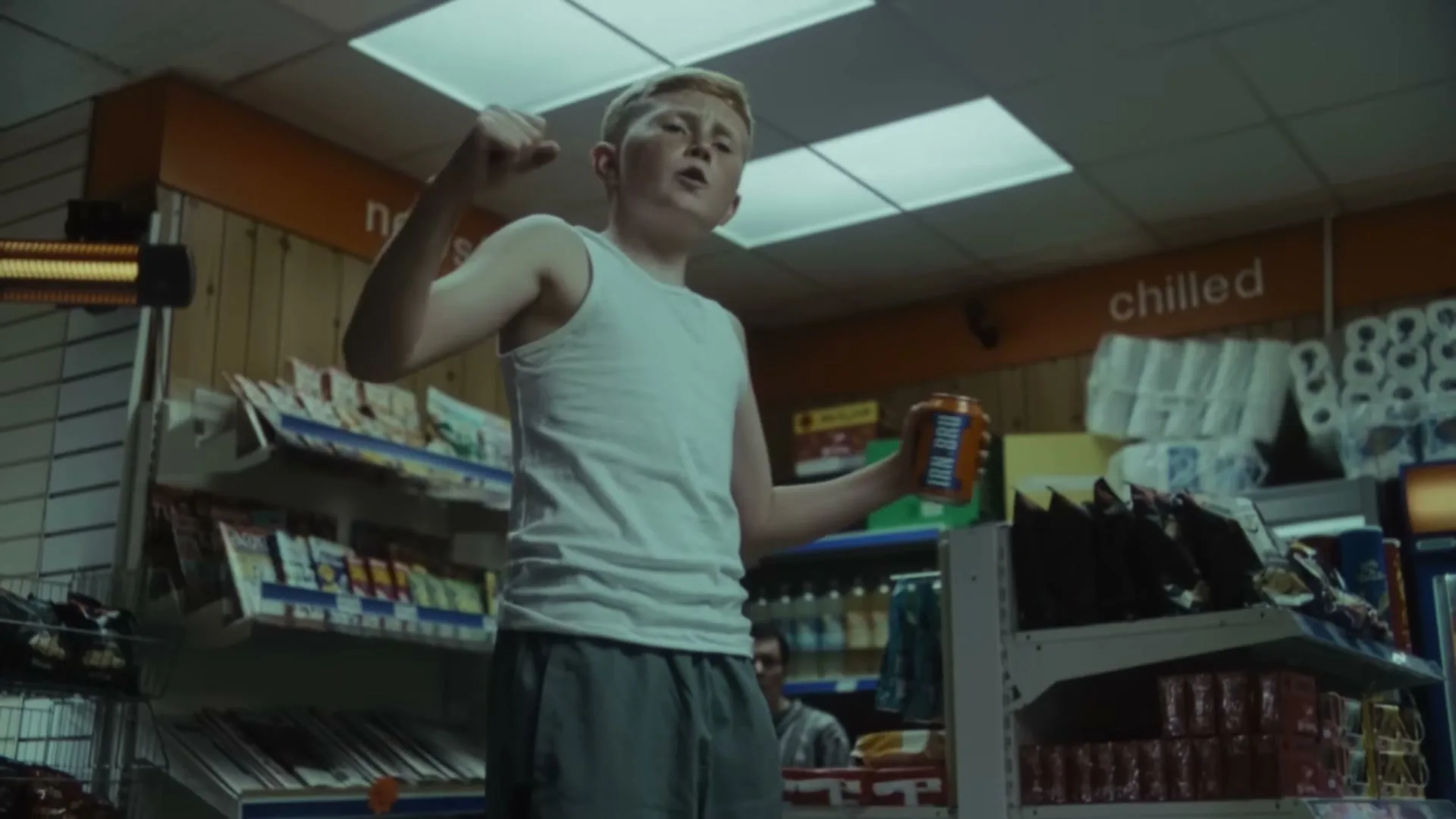 A young boy with red hair, wearing a white tank top and grey shorts, holds an IRN-BRU can and gestures with his other hand in a convenience store.