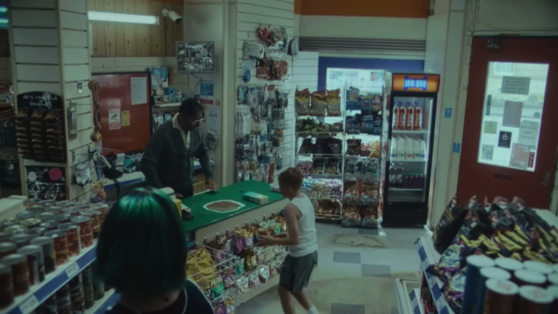 A young boy in a white tank top and grey shorts attempts to steal a bag of chips from a convenience store, while a man behind the counter watches him.