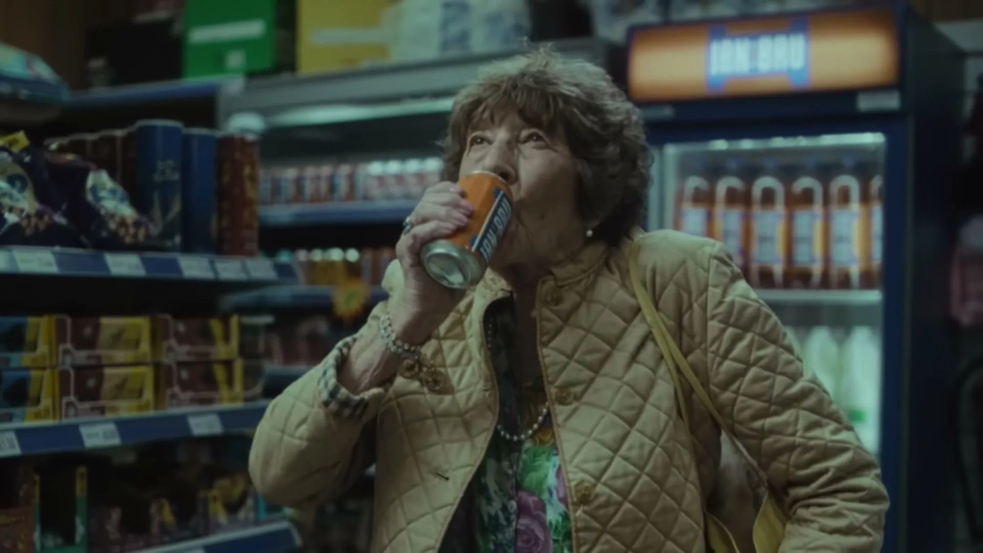 An elderly woman in a quilted jacket drinks an IRN-BRU can in a convenience store aisle.