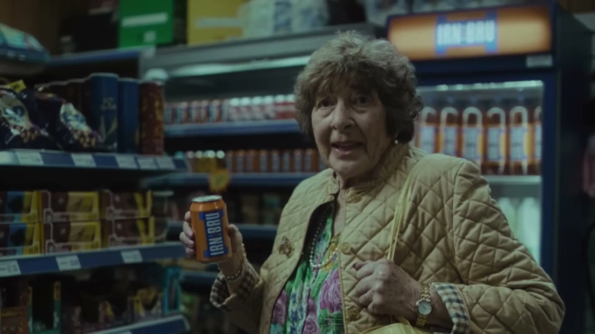 An elderly woman with brown hair, wearing a quilted jacket and floral top, holds a can of IRN-BRU in a supermarket aisle.