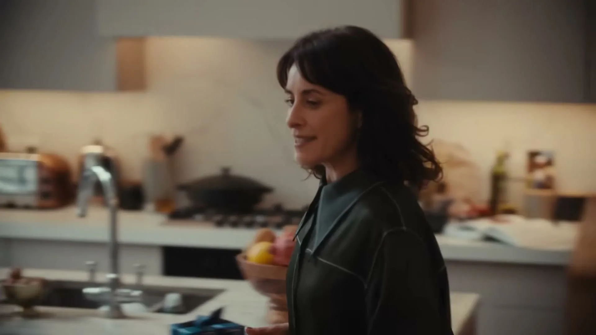 A woman with dark hair in a dark green shirt walks through a modern kitchen, gesturing with her hand as she speaks.