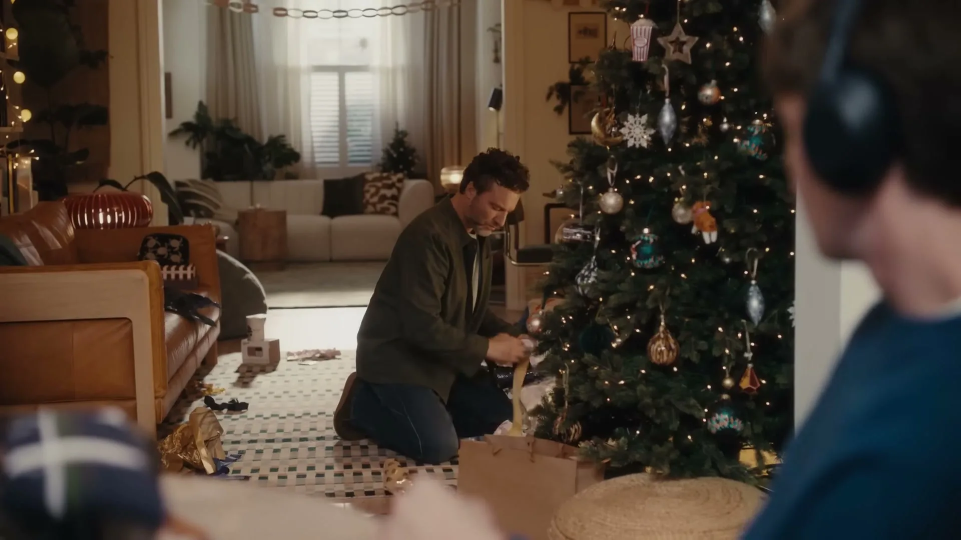 A man kneels by a Christmas tree, placing gifts underneath, while another person with headphones is seen in the foreground in a cozy living room.