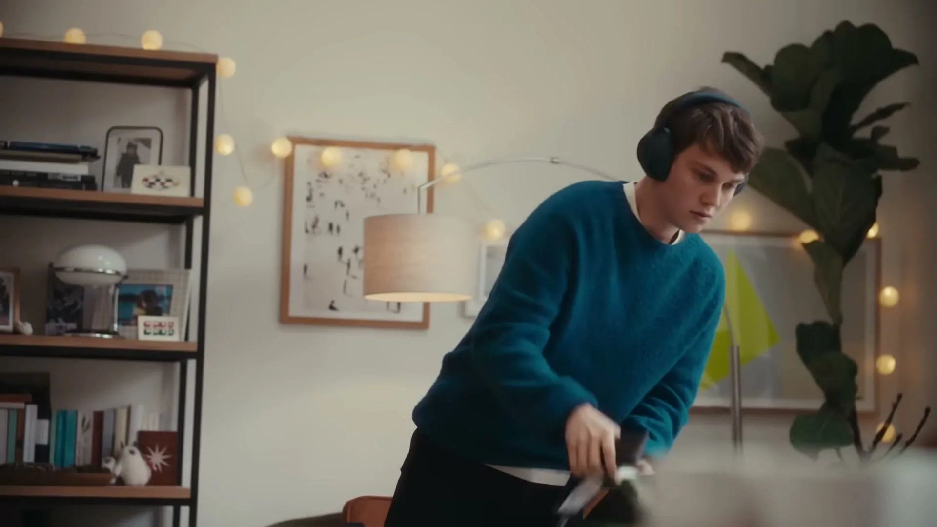A young man wearing headphones and a blue sweater leans over a table, tidying up in a cozy living room decorated with string lights.