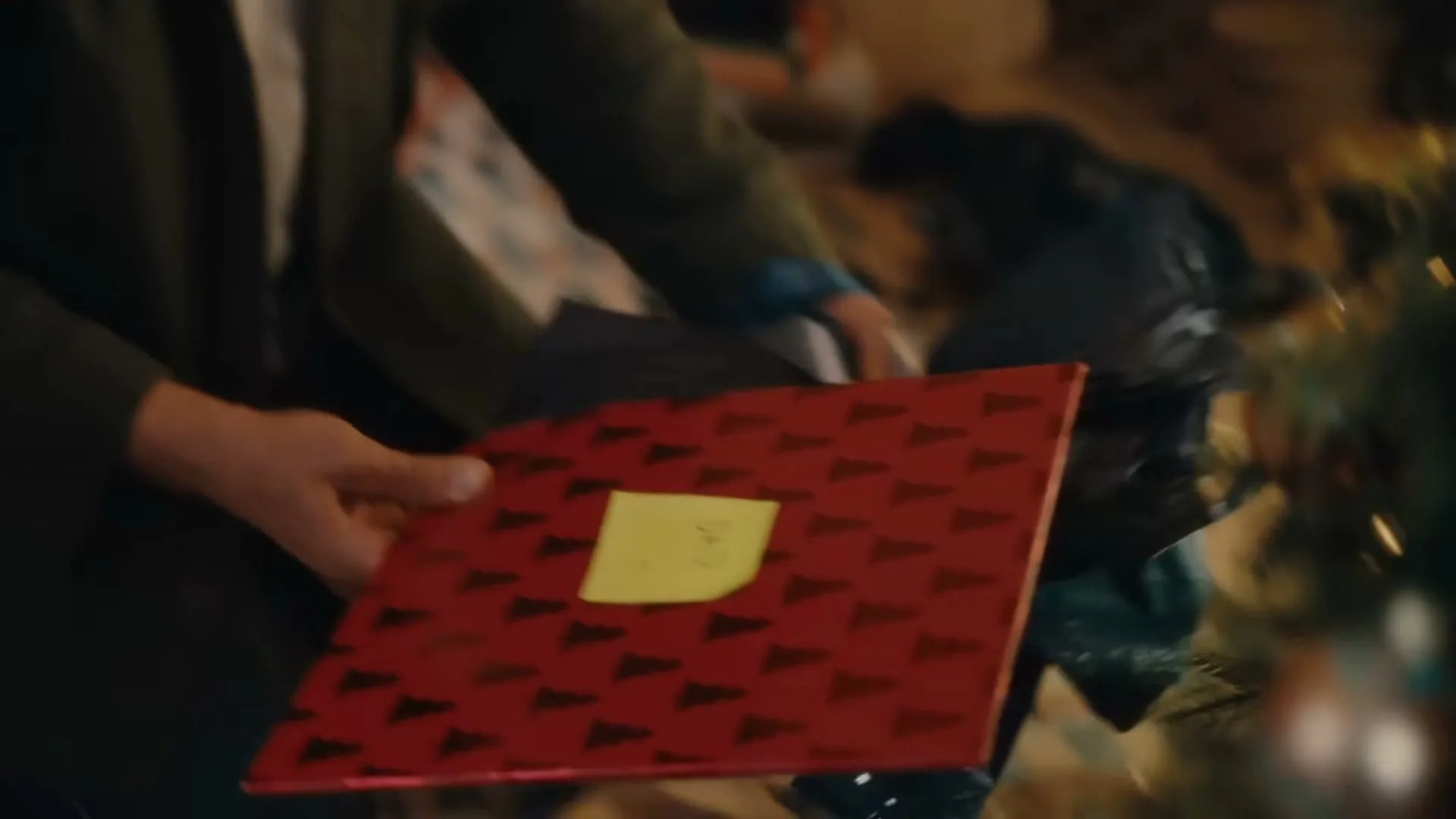 A man with a beard and dark green shirt sits on the floor, wrapping a Christmas present in a cozy living room.