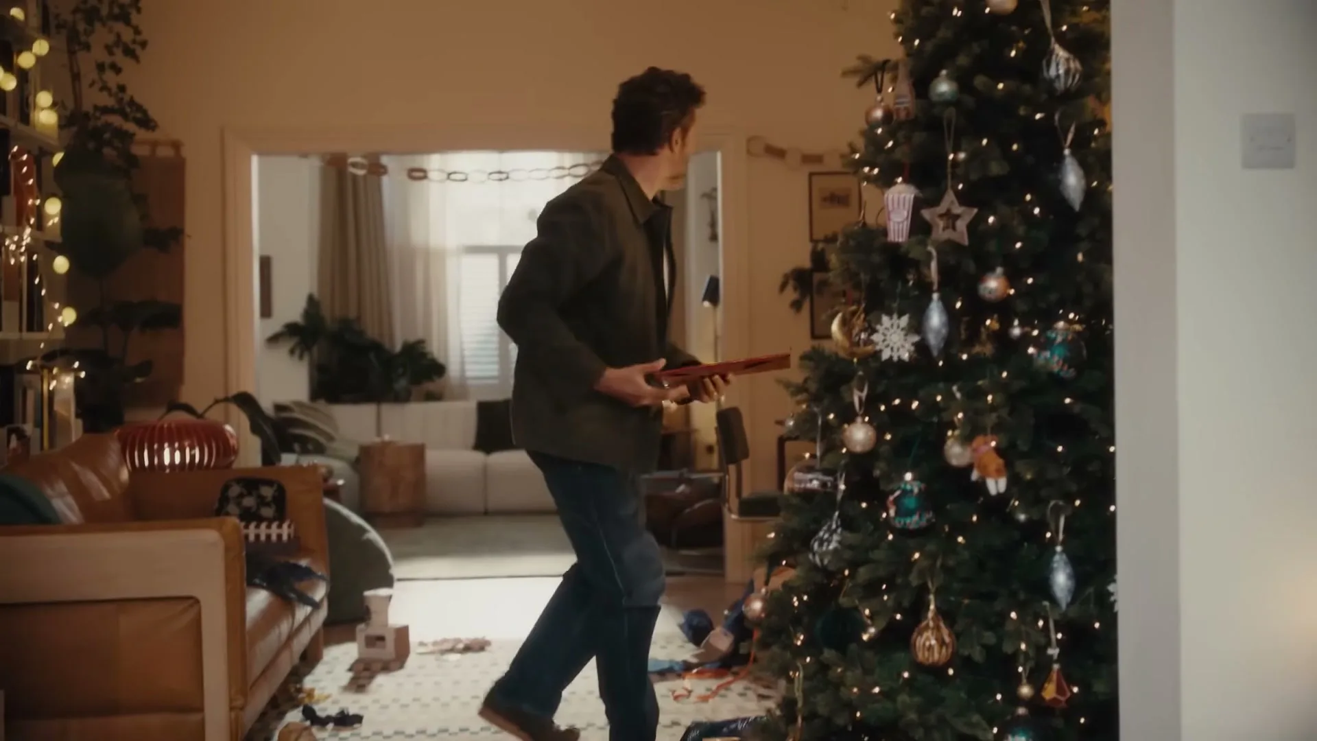A man kneels by a Christmas tree, then stands up and walks away, holding a red gift box in a cozy living room.