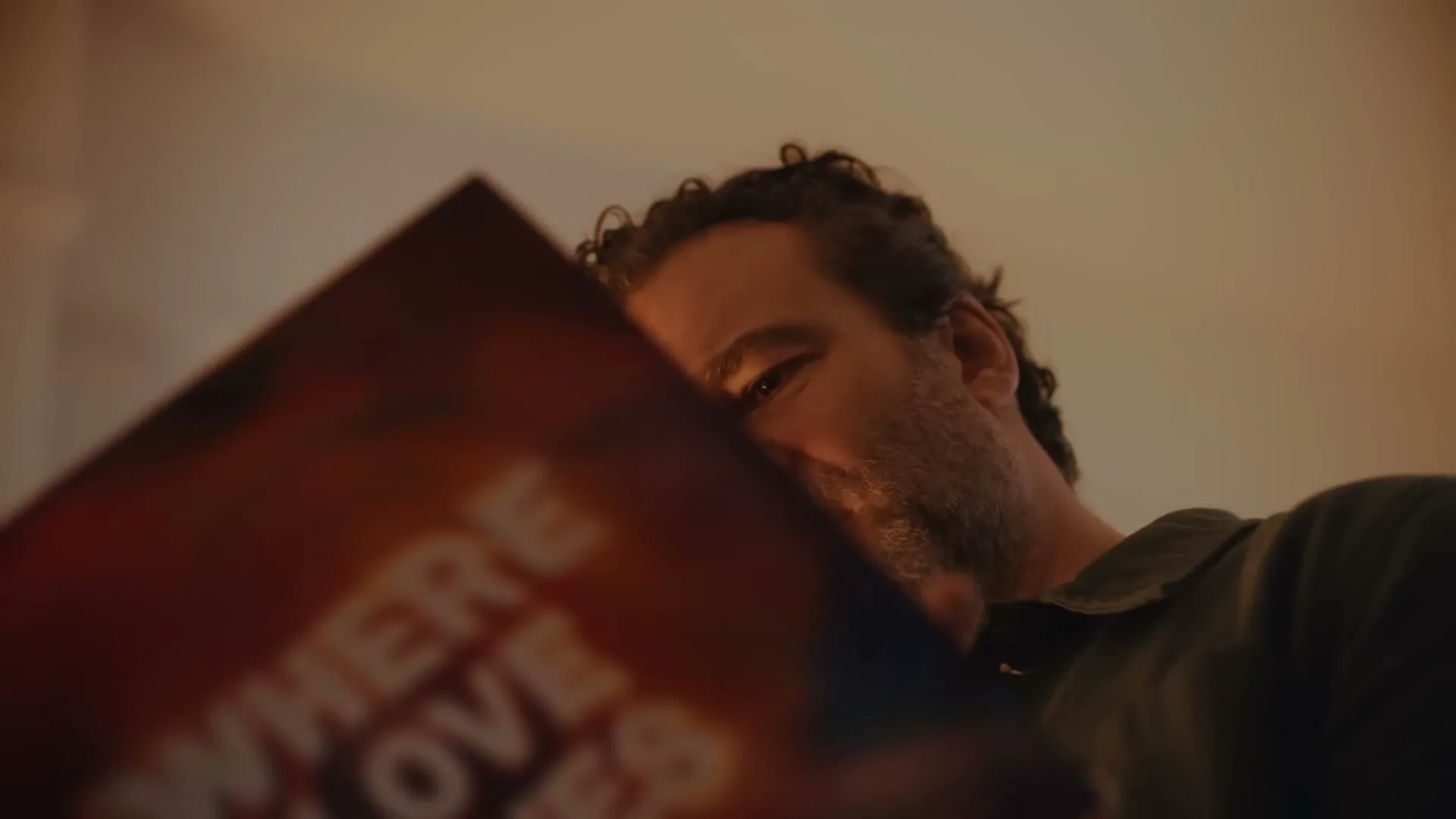 A man with curly hair and a beard looks down at a colorful book or album cover, with a slightly confused expression in a warm, dimly lit room.