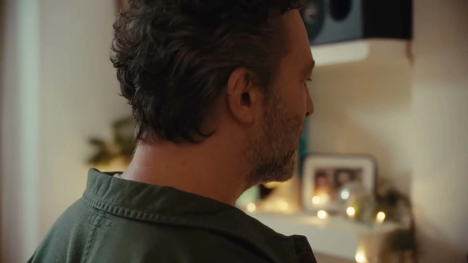 A man with a beard and dark hair, wearing a green shirt, looks down and then up, in a cozy, decorated interior.