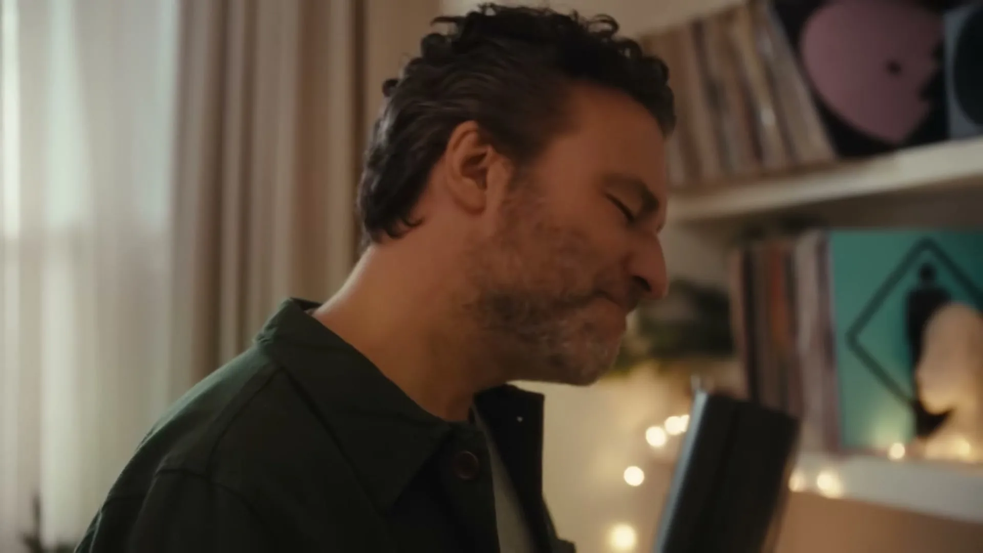A man with dark, curly hair and a beard, wearing a dark green shirt, sings into a black microphone in a cozy room with records on shelves.