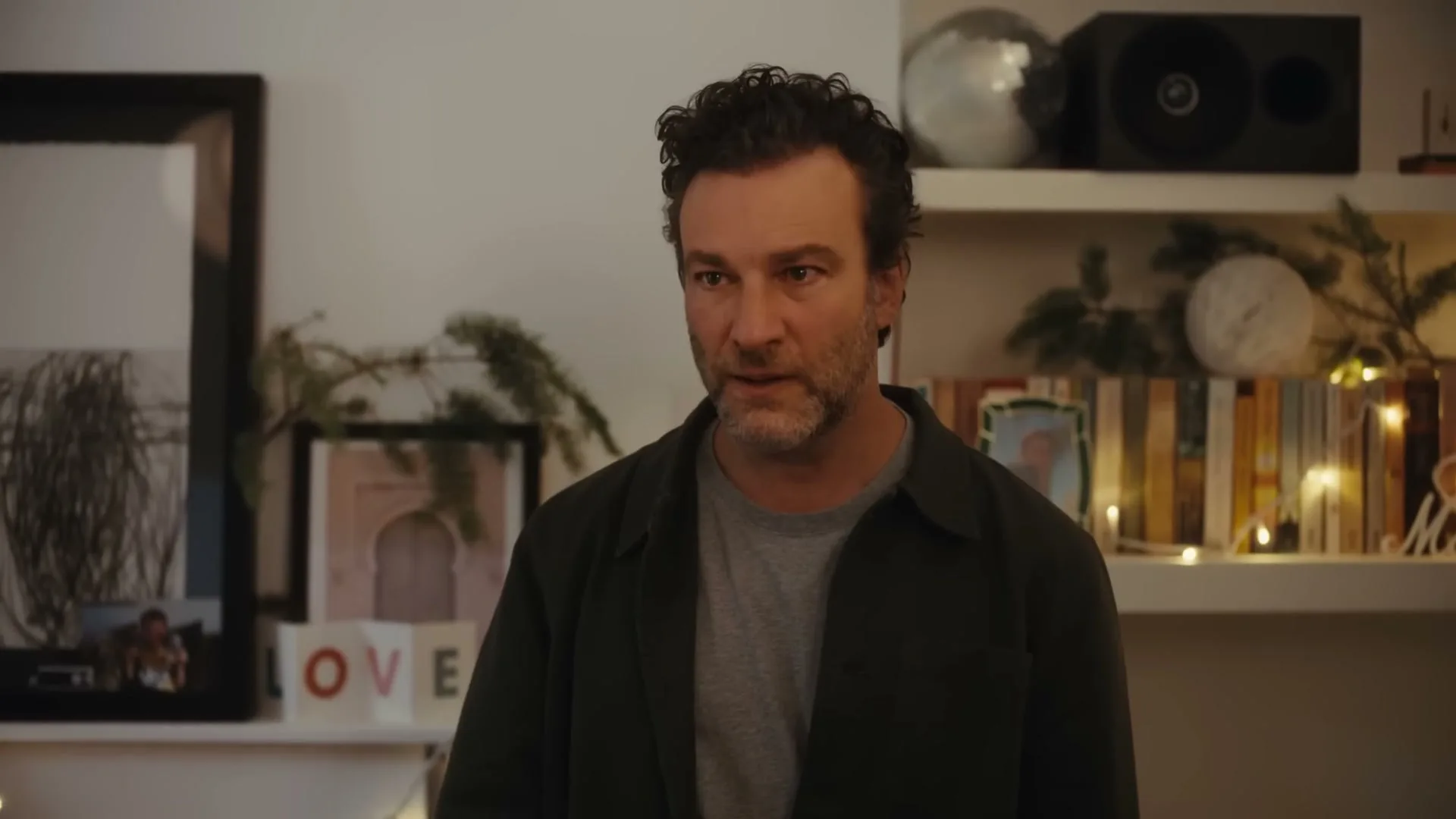 A man with dark, curly hair and a beard looks down, then up with a concerned expression in a decorated living room.