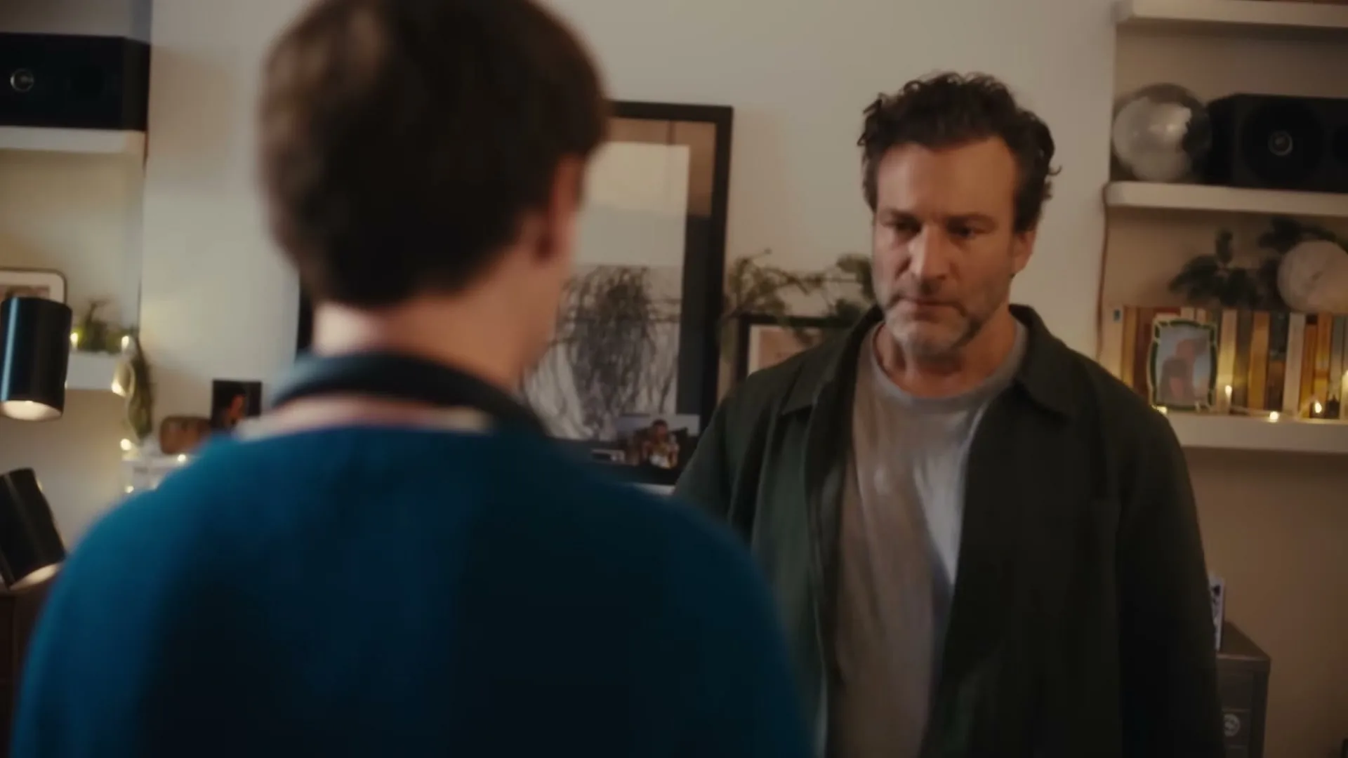 A man with dark, curly hair and a beard looks at another person, then embraces them in a warmly lit living room decorated with string lights.