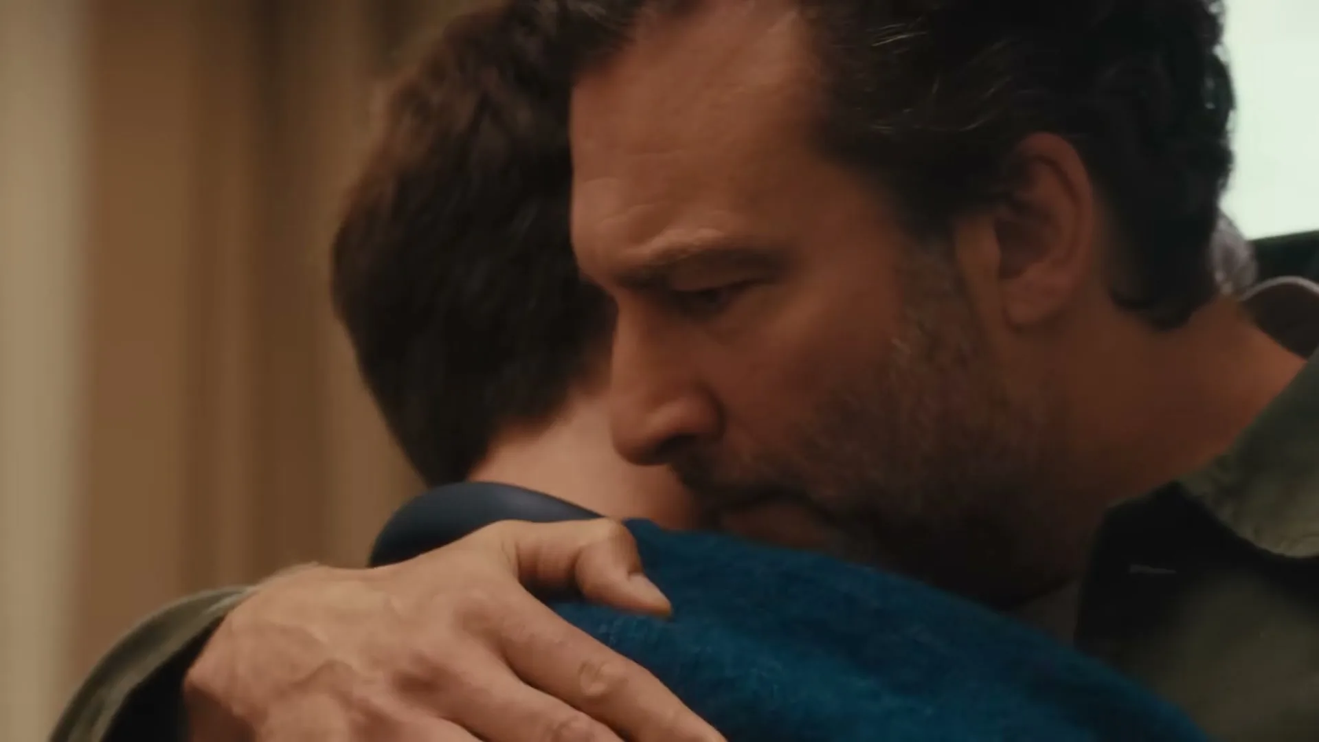 A man with a beard hugs a younger person, burying his face into their shoulder, showing affection in a warm, intimate moment indoors.