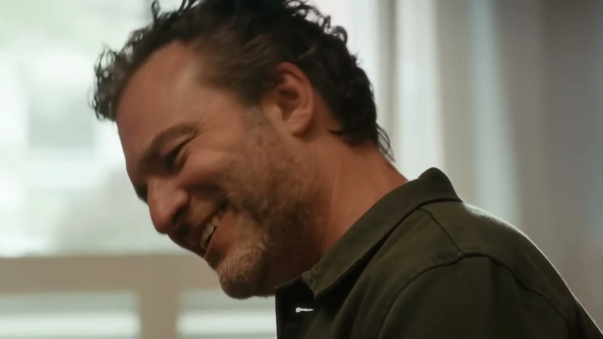 A man with dark, curly hair and a beard, wearing a green shirt, smiles and laughs while looking down and to the right in an interior setting.