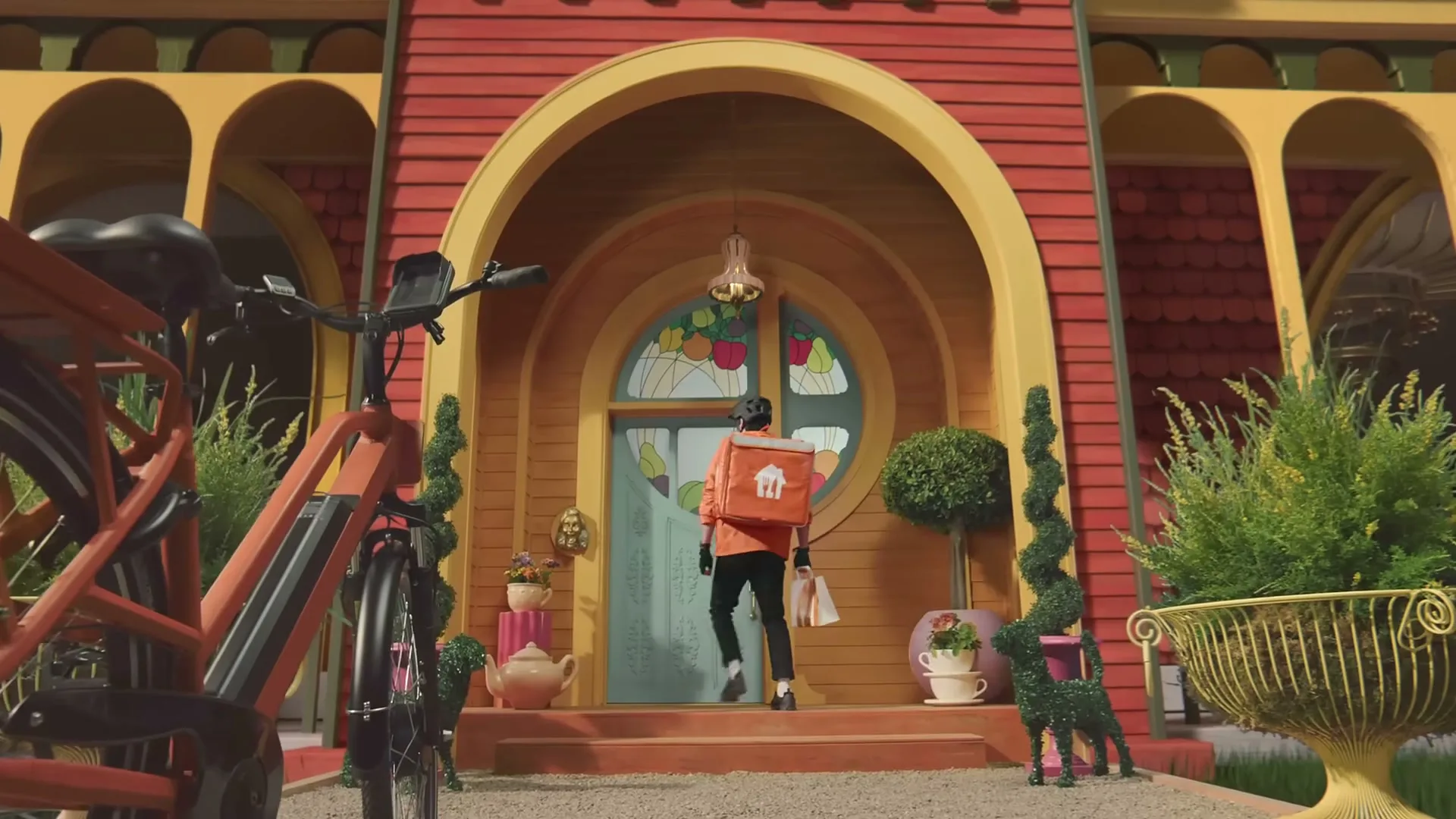 A delivery person in an orange jacket and black pants walks up the steps to a whimsical, red house with a large, arched doorway and a stained-glass window, carrying a delivery bag.