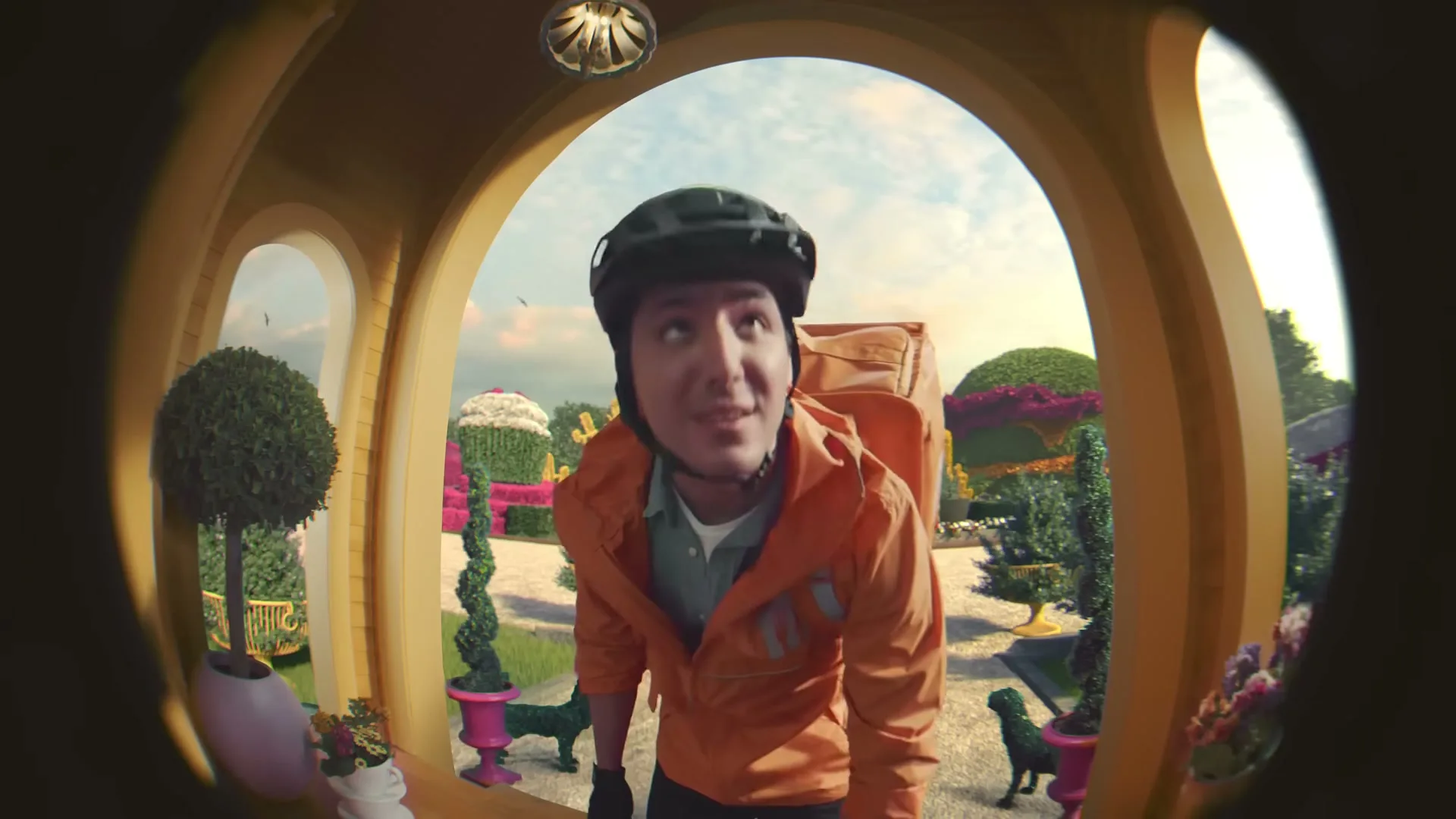 A delivery driver in an orange jacket and helmet looks into a fisheye lens, as if through a peephole, in a whimsical garden setting.