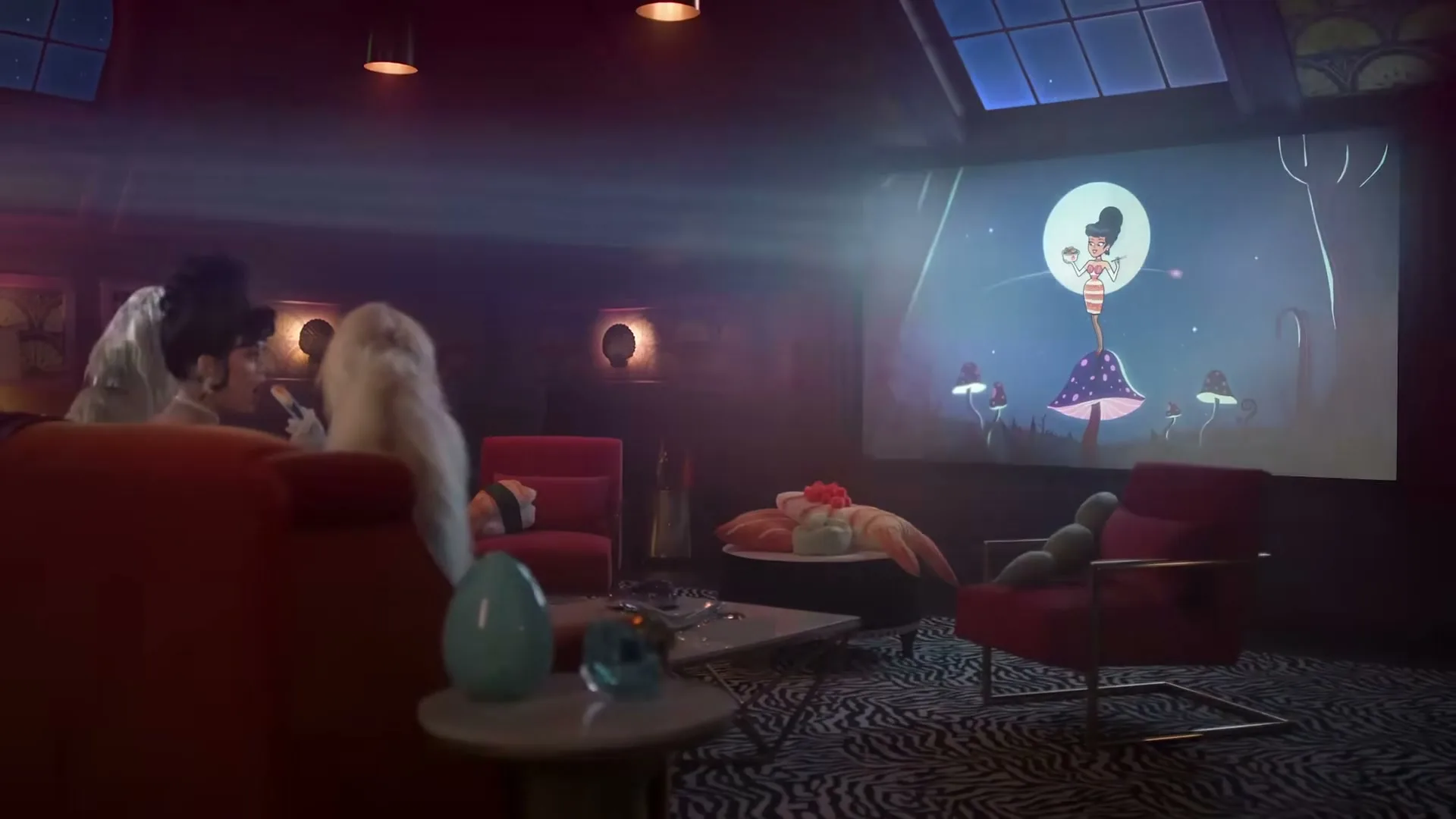 Two women in elaborate outfits watch an animated woman on a large screen in a dimly lit, stylish living room, with sushi on a coffee table.