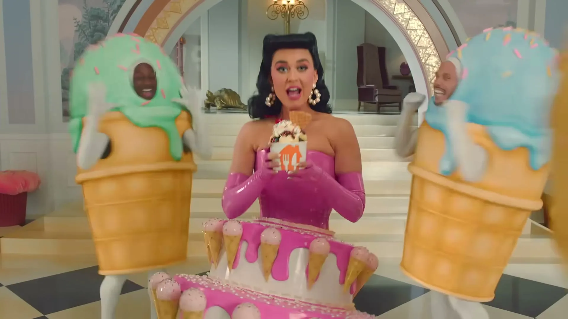 Katy Perry, dressed in a pink latex top and cake skirt, holds an ice cream sundae while surrounded by people in ice cream cone costumes in a whimsical mansion.
