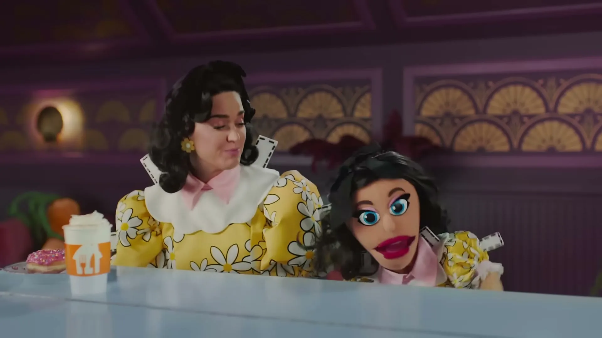 Katy Perry, dressed in a yellow daisy-print dress, sits at a diner counter with a puppet version of herself, both looking at each other.