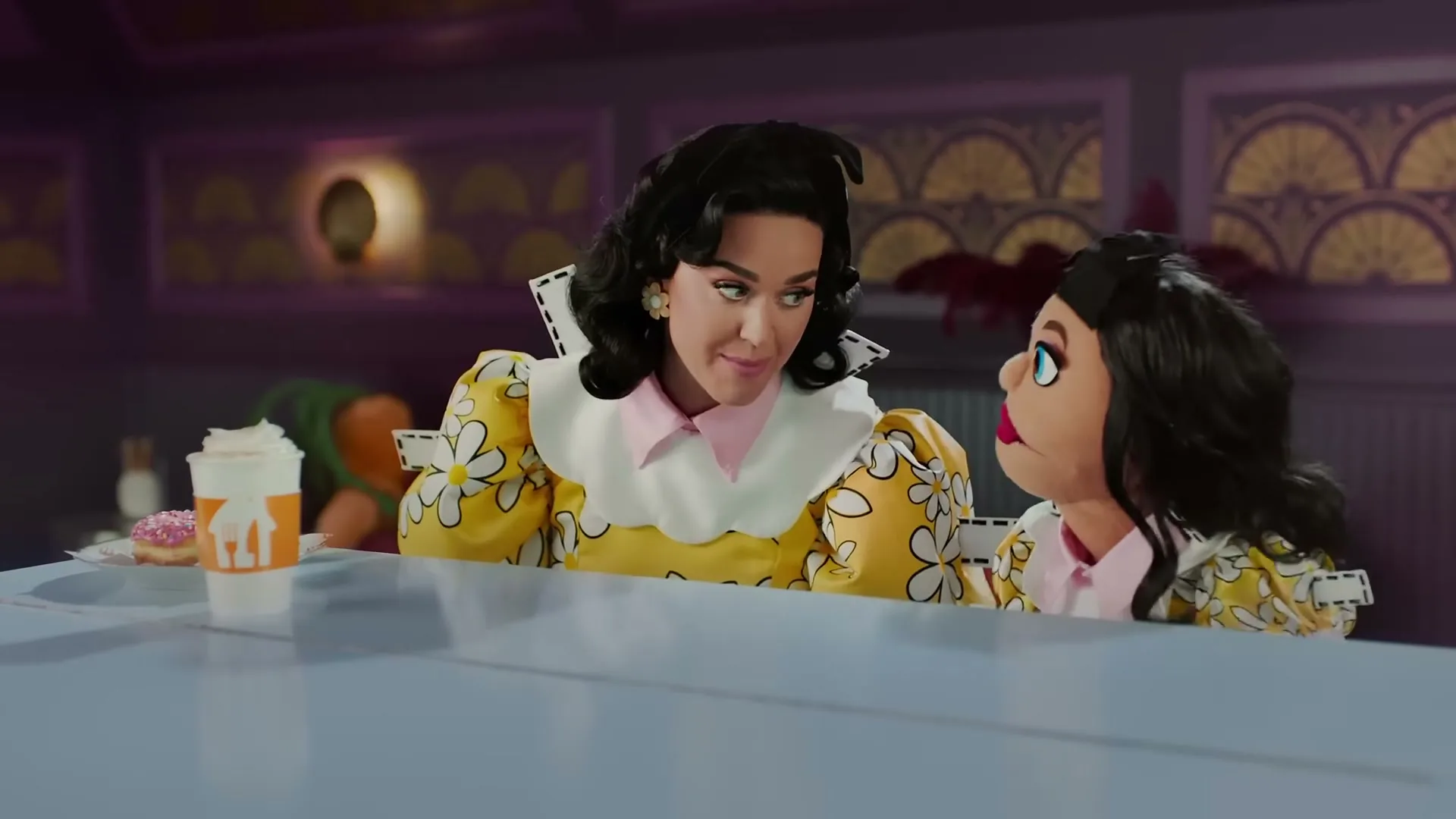 Katy Perry, dressed in a yellow floral dress, talks to a puppet version of herself at a diner counter, with a drink and donut visible.