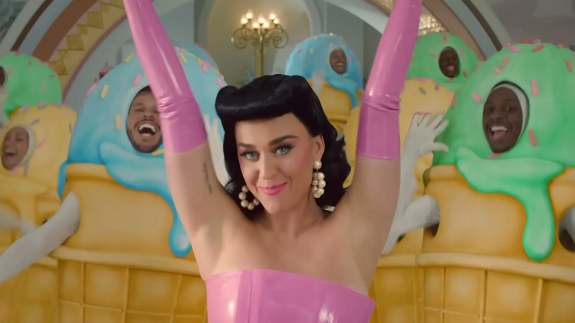 Katy Perry, dressed in a pink latex dress and long gloves, winks and raises her arms while surrounded by people in ice cream cone costumes in a whimsical room.