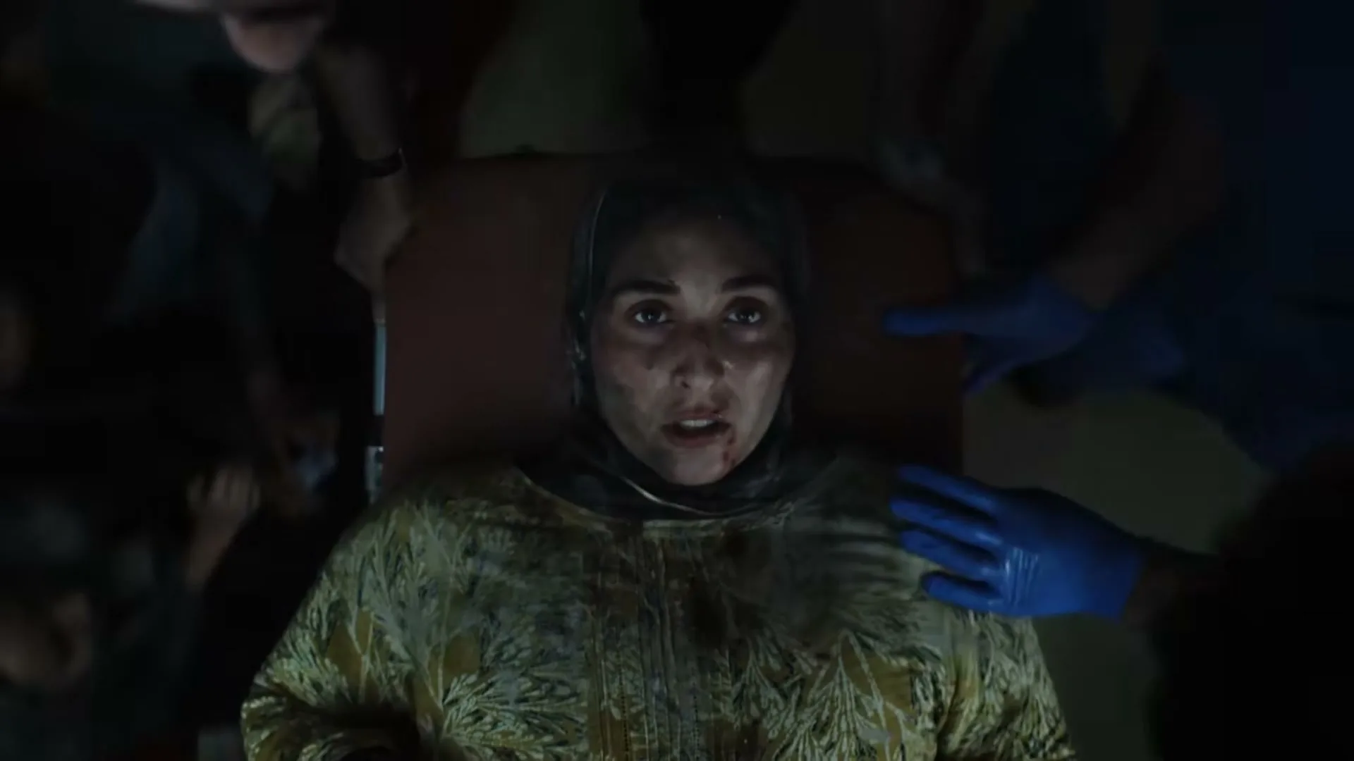 A woman with a headscarf and blood on her face lies on a stretcher, surrounded by medical personnel in a dark, chaotic environment.